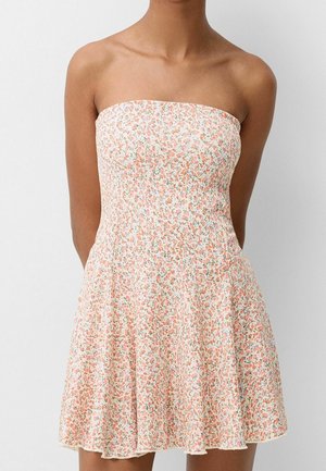 Femme portant une robe courte fleurie rose et blanche sans bretelles, debout devant un fond clair uni.