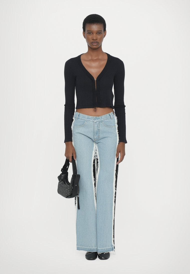 Pull cropped noir à côtes avec fermeture éclair, associé à un jean baggy bleu clair avec des bords effilochés et des franges. Tenant un sac noir à clous.