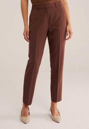 Femme portant un pantalon marron taille haute à coupe droite et des talons beiges à bout pointu, debout devant un fond uni.