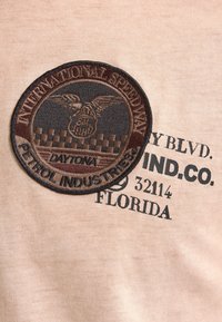 Tkanina v odstínu tan s kruhovou vyšitou nášivkou s textem "INTERNATIONAL SPEEDWAY", černobílými kostkami a detailem "DAYTONA".
