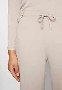 Hellbeige, gerippte Jogginghosen mit einem elastischen Kordelzug. Der Stoff ist glatt und zeigt feine Rillen. Eine Hand hält sanft die Seite der Hose.