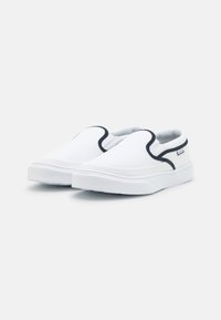 Weiße Slip-On Sneaker mit schwarzer Kontrastkante und einem kleinen Logohänger an der Seite, vor einem schlichten hellen Hintergrund präsentiert.