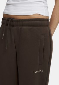 Bruna svettbyxor med resårmidja, dragsko, sidofickor och en liten logotyp "CASPARA" i ljus färg tryckt nära fållen.