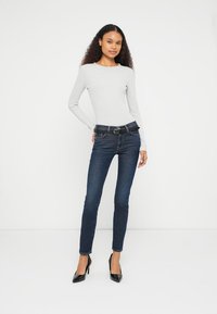 Jeans skinny azul escuro com silhueta ajustada, combinados com uma blusa de manga comprida cinza claro. O conjunto inclui um cinto preto e saltos pretos.