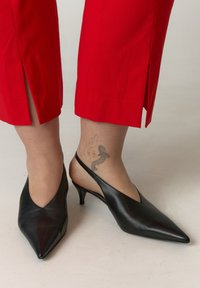 Zwarte leren slingback-hakken met spitse neus, lage kittenhakken en een V-vormige uitsnijding; gecombineerd met rode enkelhoge broek.