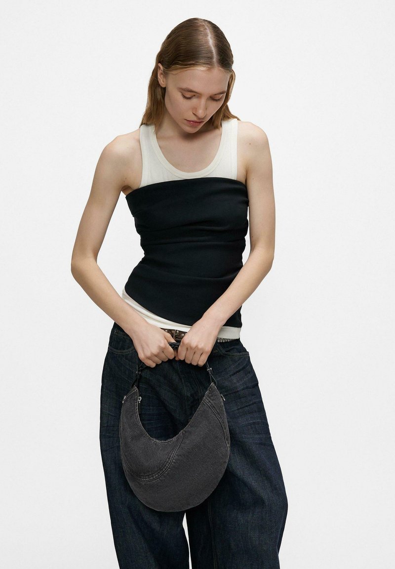 Frau mit einem zweifarbigen ärmellosen Top und dunklen Jeans mit weitem Bein, die eine schwarze, halbmondförmige Umhängetasche vor einem weißen Hintergrund hält.