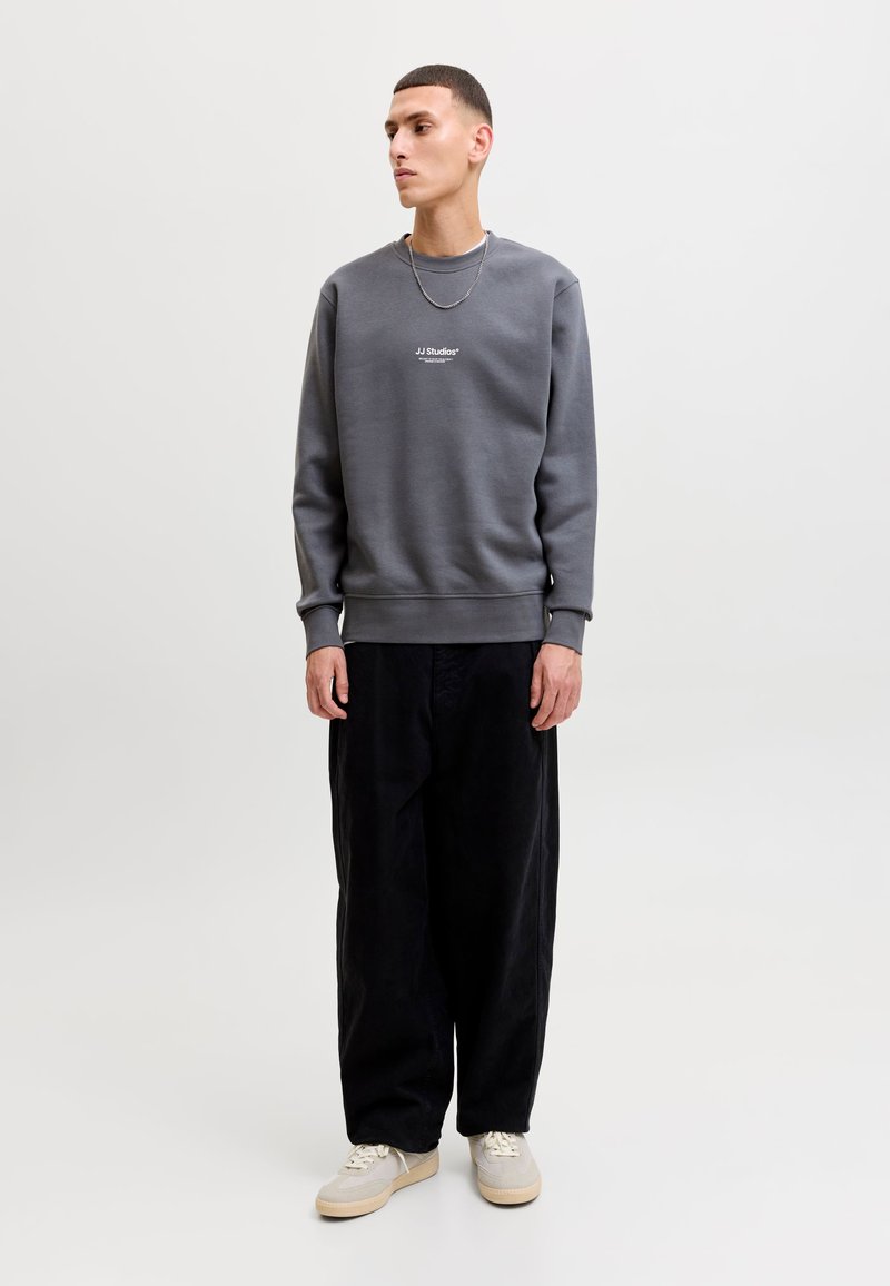 Jeune homme debout portant un sweat-shirt gris avec le texte "JJ Studios", pantalon large noir, baskets blanches et une chaîne en argent au cou.