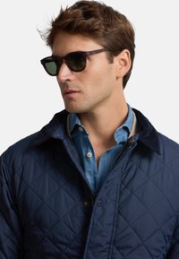Chaqueta acolchada azul marino con cuello de pana, combinada con una camisa vaquera azul claro y gafas de sol oscuras, con detalles de cremallera y botones.