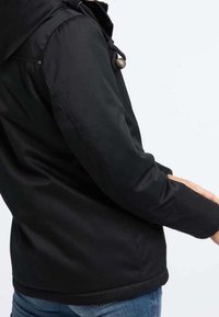 Musta vedenpitävä takki, jossa on huppu, kiristysnauha ja nappikoristukset, pehmeä tekstuuri ja istuva malli.