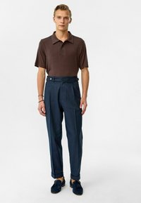 Bruine polo met kraag, gecombineerd met hoge navy broek met zijknopen. Model draagt blauwe suède instappers.