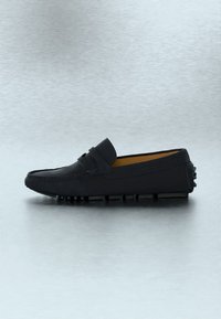 Μαύρο δερμάτινο μοκασίνι με ελαστικά καρφιά στη σόλα και λεπτομέρεια με λουράκι στο πάνω μέρος, εμφανιζόμενο σε πλευρικό προφίλ σε ανοιχτό γκρι φόντο.