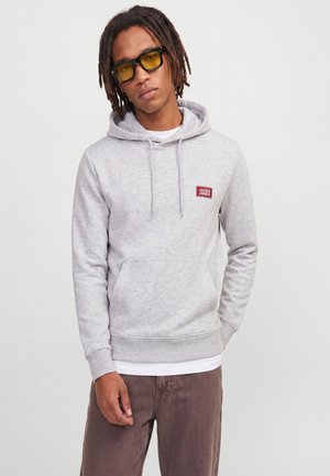 Jeune homme avec des dreadlocks portant des lunettes à verres jaunes et un sweat à capuche Jack & Jones gris clair avec un pantalon marron sur un fond uni.