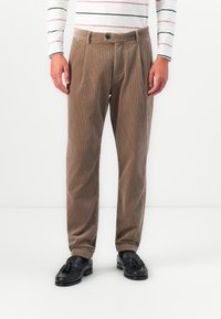 Uomo che indossa pantaloni in velluto a coste beige, mocassini neri con nappine e una camicia bianca a maniche lunghe con sottili righe orizzontali nere e rosse.