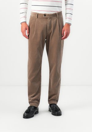 Mann trägt beigen Cordhosen, schwarze Quasten-Lederschuhe und ein weißes Langarmhemd mit schmalen schwarzen und roten horizontalen Streifen.