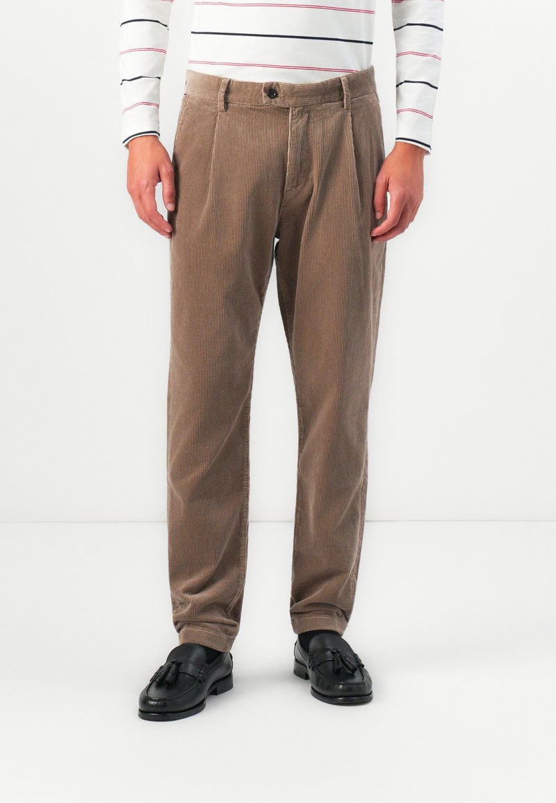 Uomo che indossa pantaloni in velluto a coste beige, mocassini neri con nappine e una camicia bianca a maniche lunghe con sottili righe orizzontali nere e rosse.