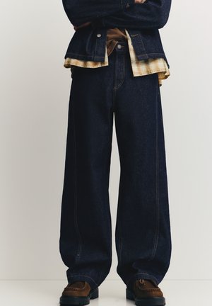 Sötét farmer széles szárú nadrágok narancssárga varrással, kockás inggel és barna cipővel párosítva. Az anyag masszívnak és texturáltnak tűnik.