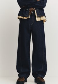 Temno modre široke kavbojke iz jeansa z oranžnimi šivi, kombinirane s karo srajco in rjavimi čevlji. Tkanina je videti trdna in teksturirana.