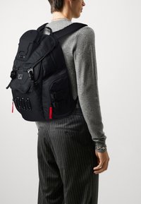 Black backpack with multiple compartments, adjustable straps, and textured fabric. Features red zipper pulls and a logo patch.
