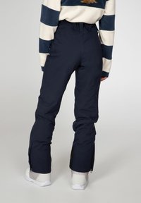 Pantalons de ski imperméables en bleu marine foncé avec une taille haute, fermetures éclair sur les côtés aux poignets, associés à des chaussures blanches et un haut rayé multicolore.