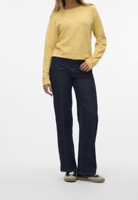 Pull jaune tricoté avec des poignets côtelés, associé à un jean évasé bleu foncé. Les chaussures présentent un design noir et beige avec des lacets.
