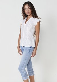 Blouse en coton blanc avec broderies florales, manches à volants et devant boutonné. Associée à un jean cropped bleu clair avec une couture décorative.