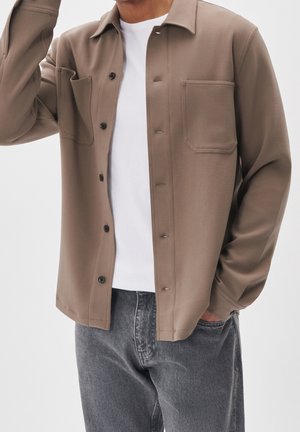 Homme portant une chemise taupe boutonnée par-dessus un t-shirt blanc avec des boutons noirs, une main dans la poche d'un jean gris.
