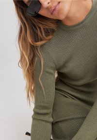 Robe en maille côtelée olive avec manches longues et encolure ronde, présentant un tissu texturé et une coupe près du corps. Lunettes de soleil noires portées.