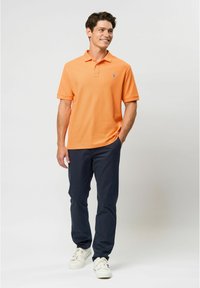 Orange polo shirt with short sleeves and ribbed collar, featuring a light blue logo. Worn with navy pants and white sneakers.