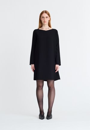 Frau mit glatten roten Haaren trägt ein schlichtes schwarzes langärmliges Kleid, schwarze transparente Strumpfhosen, schwarze High Heels und eine silberne Halskette, steht vor weißem Hintergrund.
