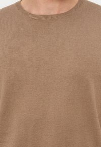 Κοντινό πλάνο ενός ατόμου που φορά ένα μονόχρωμο, ανοιχτό καφέ πλεκτό πουλόβερ με στρογγυλή λαιμόκοψη σε λευκό φόντο.