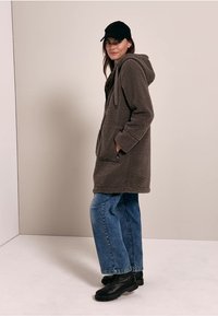 Hoodie marron surdimensionnée, doublée en polaire, avec une poche frontale et un cordon de serrage, portée sur un jean en denim bleu ample, associée à des bottes noires.