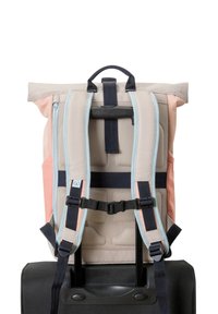 Le sac à dos présente des bretelles rembourrées dans des couleurs pastel, en tissu beige, avec une texture en mesh au dos, et une poignée. Il s'attache à une valise noire.