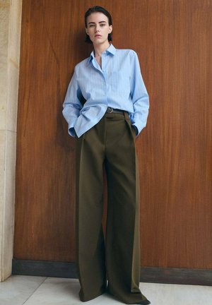 Persoană stând cu spatele la un perete din panouri de lemn, purtând o cămașă albastră largă cu nasturi și pantaloni maro cu talie înaltă, largi, cu mâinile în buzunare.