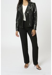 Person i svart läderjacka, vit topp, svarta byxor och svarta klacksandaler mot en enkel vit bakgrund.