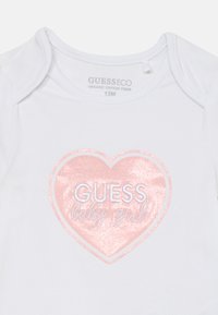 Bijela organic pamuk majica sa dugim rukavima koja ima dizajn ružičaste srca s glitterom i natpisom "GUESS baby girl" u plavoj boji. Veličina 12M.
