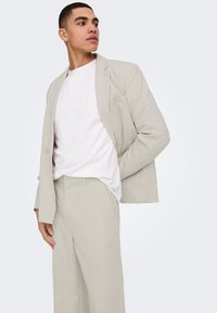 Traje beige claro con blazer ajustado y pantalones de pierna ancha, combinado con una camiseta blanca de cuello redondo. Tejido suave y diseño minimalista.