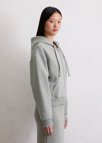 Haut de sweat à capuche zippé gris clair avec une capuche à cordon de serrage, des poignets côtelés et une poche kangourou, fabriqué dans un tissu doux et texturé.