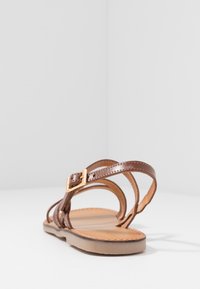 Sandalia de cuero marrón con una correa ajustable con hebilla, suela plana y diseño recortado. Presenta costuras mínimas y un acabado suave.