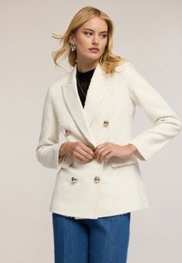 Biała, teksturowana, dwurzędowa marynarka z złotymi guzikami, posiadająca klapy i boczne kieszenie. W komplecie z niebieskimi dżinsami.