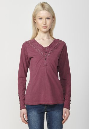 Chemise à manches longues bordeaux avec un col en V, des détails en dentelle le long de l'encolure et des accents de boutons aux poignets.