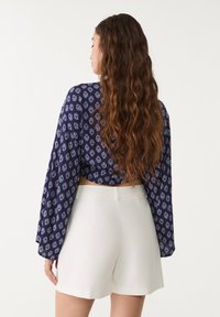 Top court bleu marine avec un motif floral blanc, manches longues évasées, associé à un short blanc taille haute, texture de tissu lisse.