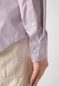 Hellpurpurnes Hemd mit einem kleinen geometrischen Muster, mit geknöpften Manschetten und glattem Stoff. Über einer hellen Hose getragen.