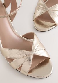 Goldfarbene, metallische High-Heel-Sandalen mit gekreuzten Riemen, offenem Zehenbereich, glatter Oberfläche und hellbrauner Innensohle. Flache schwarze Laufsohle.