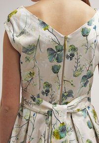 Kukkamekko, jossa on kevyt kuvioitu kangas, syvällä V-muotoisella selällä, piilotettu vetoketju ja sidottava vyö; värit sisältävät vihreitä ja keltaisia korostuksia.