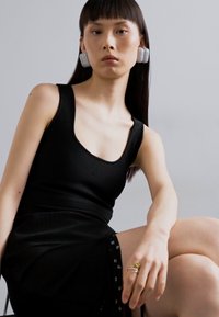 Top negru fără mâneci cu decolteu rotunjit, asortat cu o fustă neagră ce are accente cu nasturi pe lateral; modelul poartă cercei mari cu textură.