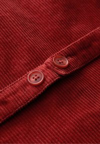 Tissu en velours côtelé rouge avec une texture côtelée et deux boutons assortis à quatre trous. Le tissu a une apparence douce et légèrement duveteuse.