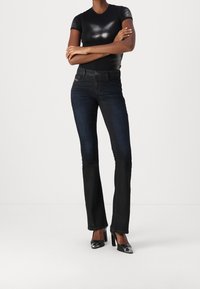 Nainen, jolla on päällään musta lyhythihainen paita, tummat istuvat bootcut-farkut ja mustat korkokengät, joissa on hopeiset kärjet, kädet ristissä.