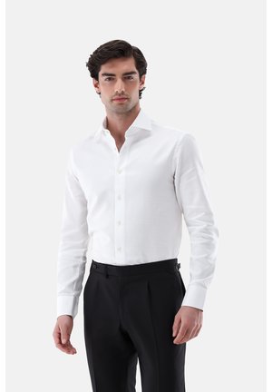 Junger Mann mit dunklem Haar, der ein weißes Hemd mit Knöpfen und schwarze Hose trägt und vor einem schlichten hellen Hintergrund steht.
