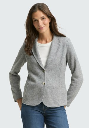 Vrouw draagt een lichtgrijs vest met knopen over een wit topje en blauwe spijkerbroek, poseert met handen in de zakken tegen een effen achtergrond.