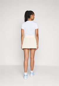 Camicia bianca a maniche corte abbinata a una gonna plissettata beige chiaro. Il modello indossa scarpe da ginnastica bianche con dettagli blu e calzini alla caviglia.